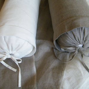 May include: Two cylindrical pillows, one white and one brown, are made of linen fabric. Both pillows have a simple, gathered design and are tied with matching fabric ribbons.