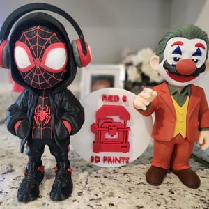 May include: Two figurines: a Miles Morales Spider-Man with headphones and a Joker-Mario hybrid. The Spider-Man figure is black and red, while the Joker-Mario figure is in a red suit with green hair. A white disc with "RED 6 3D PRINTS" in red is between them.