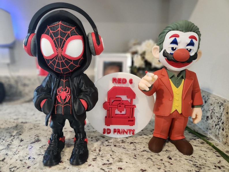May include: Two figurines: a Miles Morales Spider-Man with headphones and a Joker-Mario hybrid. The Spider-Man figure is black and red, while the Joker-Mario figure is in a red suit with green hair. A white disc with "RED 6 3D PRINTS" in red is between them.