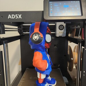 May include: A 3D-printed figure of a superhero wearing a blue hoodie, orange sleeves, and blue sneakers. The figure has a red and black mask and headphones. The figure is on a 3D printer with a digital display showing settings.