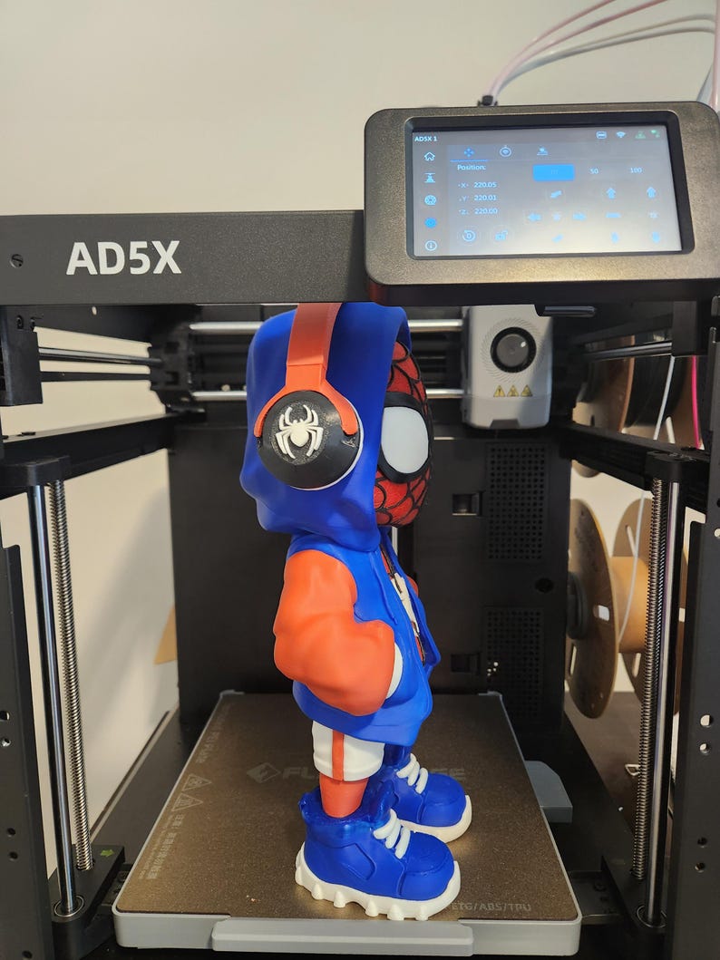 May include: A 3D-printed figure of a superhero wearing a blue hoodie, orange sleeves, and blue sneakers. The figure has a red and black mask and headphones. The figure is on a 3D printer with a digital display showing settings.