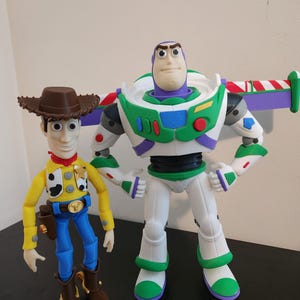 May include: A plastic action figure of a cowboy wearing a brown hat, yellow shirt, and blue trousers standing next to a plastic action figure of a space ranger with a purple hood, white and green suit, and retractable wings with red and white stripes.