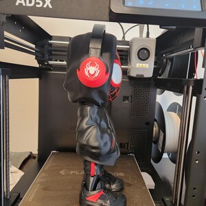 May include: A 3D-printed figure of a character wearing a black hoodie, sneakers, and headphones with a red spider emblem. The figure is on a 3D printer with the text "AD5X" on the top.