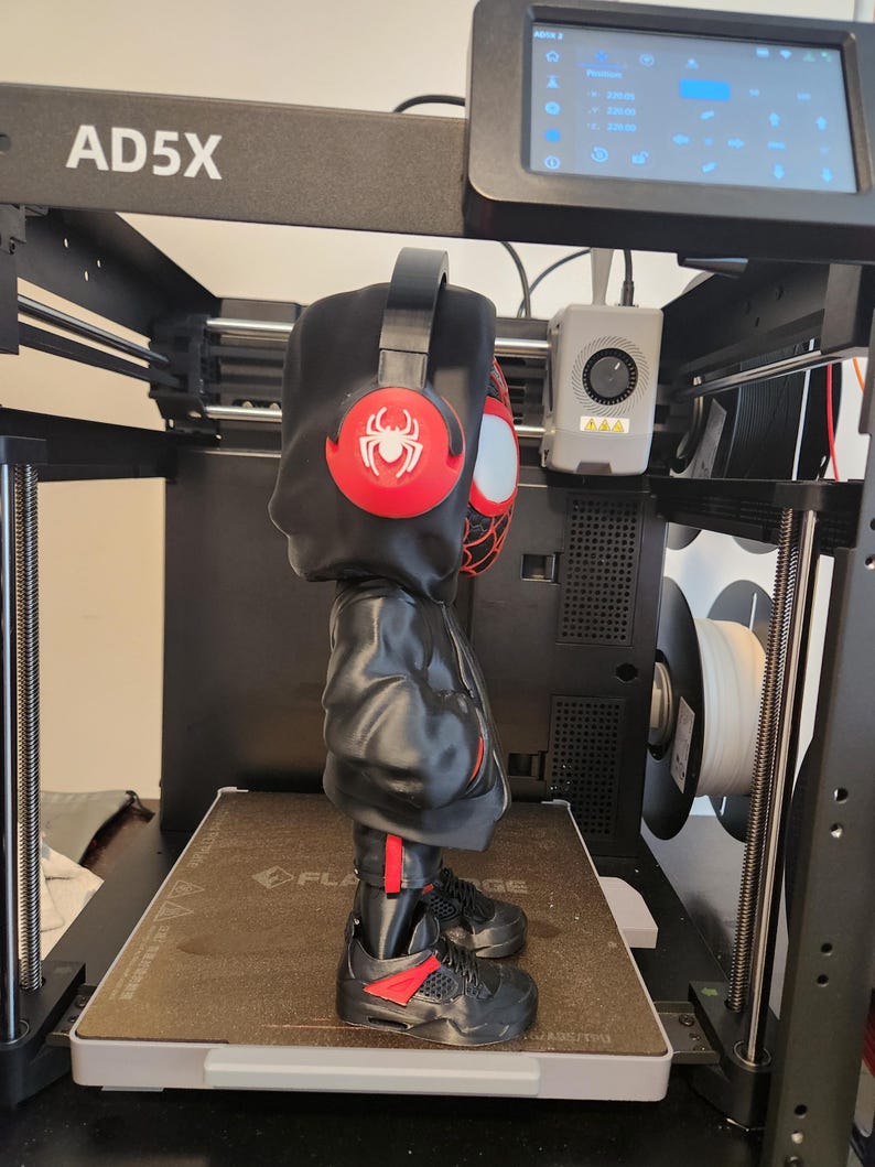 May include: A 3D-printed figure of a character wearing a black hoodie, sneakers, and headphones with a red spider emblem. The figure is on a 3D printer with the text "AD5X" on the top.