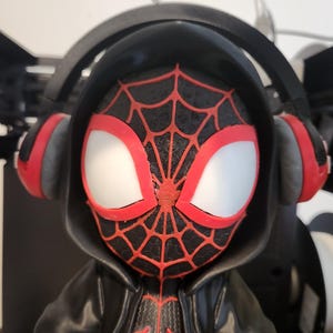 May include: A figurine of a superhero with a black and red spiderweb design on the face and chest. The figure wears a black hooded jacket and headphones with red accents. The eyes are white with red outlines.