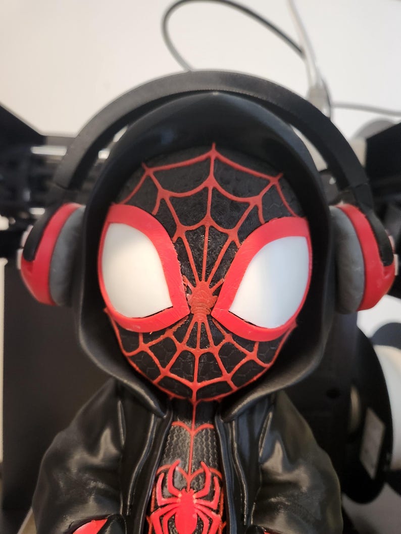 May include: A figurine of a superhero with a black and red spiderweb design on the face and chest. The figure wears a black hooded jacket and headphones with red accents. The eyes are white with red outlines.