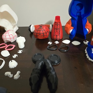May include: A collection of disassembled Spider-Man and Spider-Gwen action figure parts. The parts include a red and black Spider-Man mask, a red and black torso, a blue and orange hooded jacket, and various white and pink accessories.