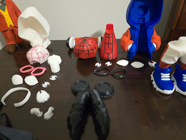 May include: A collection of disassembled Spider-Man and Spider-Gwen action figure parts. The parts include a red and black Spider-Man mask, a red and black torso, a blue and orange hooded jacket, and various white and pink accessories.