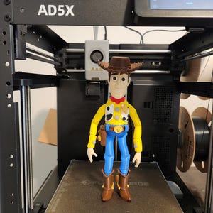 May include: A 3D-printed figurine of a cowboy character standing on the print bed of an AD5X 3D printer. The figure wears a brown hat, yellow shirt with black and white cow-print vest, red neckerchief, blue trousers, and brown boots with a holster.