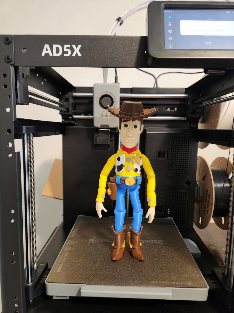 May include: A 3D-printed figurine of a cowboy character standing on the print bed of an AD5X 3D printer. The figure wears a brown hat, yellow shirt with black and white cow-print vest, red neckerchief, blue trousers, and brown boots with a holster.