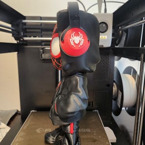 May include: A black figurine of a character wearing a black hoodie, red and black headphones, and black trainers with red accents. The headphones have a red circle with a white spider logo. The figurine is standing on a brown surface.
