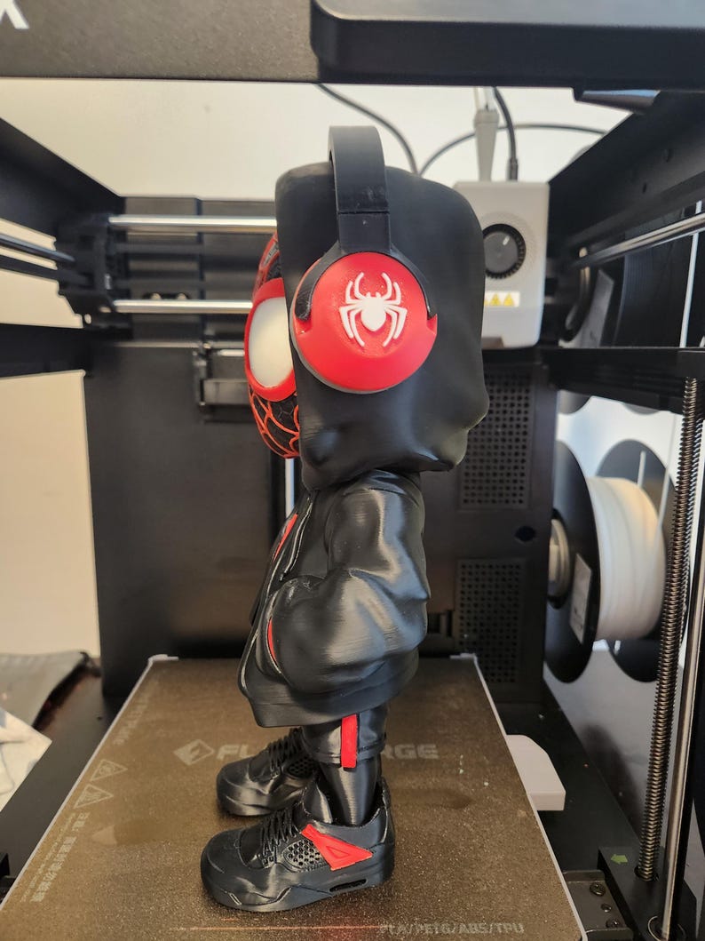 May include: A black figurine of a character wearing a black hoodie, red and black headphones, and black trainers with red accents. The headphones have a red circle with a white spider logo. The figurine is standing on a brown surface.