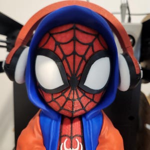May include: A 3D-printed figurine of Spider-Man wearing a blue hoodie and orange jacket. The figure has white eyes and a red spider-web design on the face. The figure is wearing red headphones with black and white accents. A white spider emblem is on the chest.
