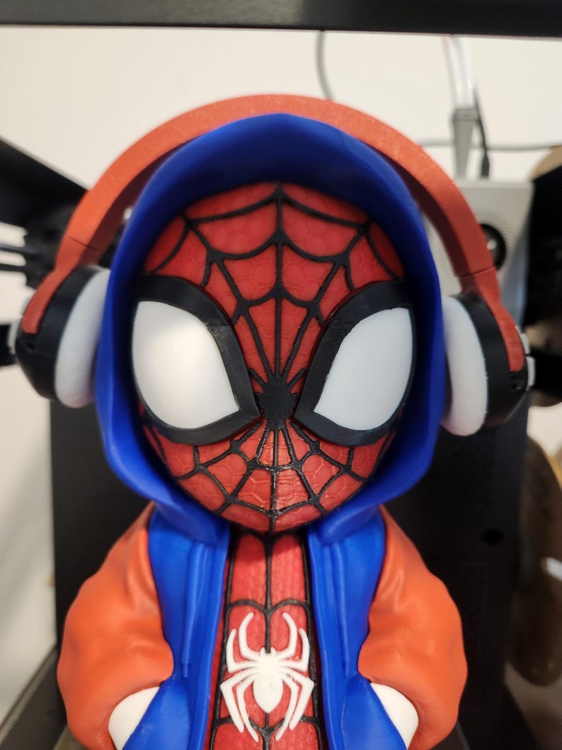 May include: A 3D-printed figurine of Spider-Man wearing a blue hoodie and orange jacket. The figure has white eyes and a red spider-web design on the face. The figure is wearing red headphones with black and white accents. A white spider emblem is on the chest.