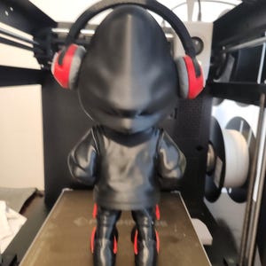 May include: A black figurine wearing a black jacket, trousers, and red-accented headphones. The figurine is standing on a brown surface, with a 3D printer in the background.