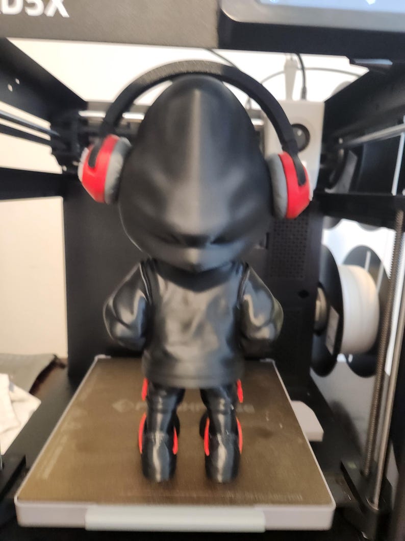 May include: A black figurine wearing a black jacket, trousers, and red-accented headphones. The figurine is standing on a brown surface, with a 3D printer in the background.
