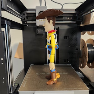 May include: A plastic figurine of a cowboy character standing on the print bed of a 3D printer. The figure has a brown hat, yellow long-sleeved shirt, white vest with black cow spots, blue trousers, and brown boots with gold-coloured spurs on the heels.