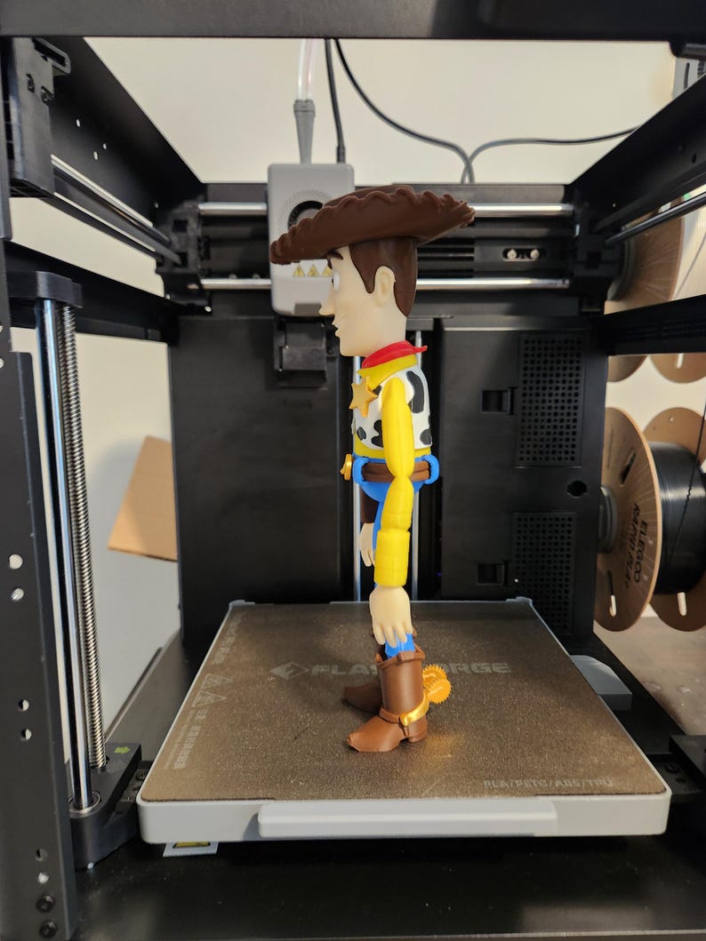 May include: A plastic figurine of a cowboy character standing on the print bed of a 3D printer. The figure has a brown hat, yellow long-sleeved shirt, white vest with black cow spots, blue trousers, and brown boots with gold-coloured spurs on the heels.