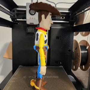 May include: A plastic action figure of a cowboy character wearing a brown hat, a yellow long-sleeved shirt with a black and white cow-print vest, a red neckerchief, blue trousers, and brown boots with yellow spurs, standing on a 3D printer bed.