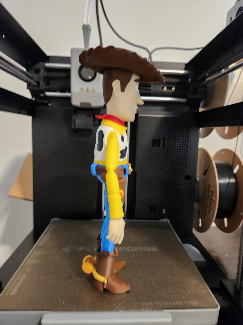May include: A plastic action figure of a cowboy character wearing a brown hat, a yellow long-sleeved shirt with a black and white cow-print vest, a red neckerchief, blue trousers, and brown boots with yellow spurs, standing on a 3D printer bed.