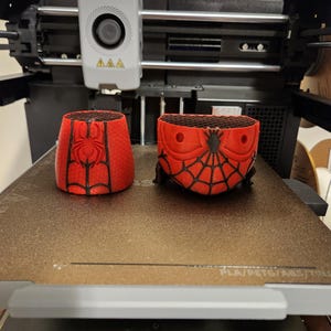 May include: Two red and black 3D-printed objects, possibly containers, featuring a Spider-Man design. The objects are on a 3D printer bed. The printer has a gray head with a circular element and three yellow triangles.