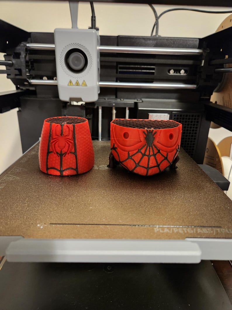 May include: Two red and black 3D-printed objects, possibly containers, featuring a Spider-Man design. The objects are on a 3D printer bed. The printer has a gray head with a circular element and three yellow triangles.
