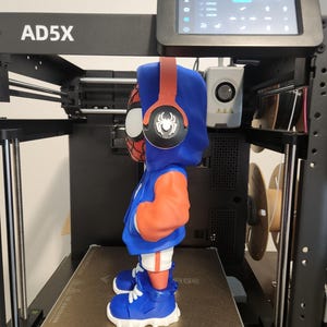 May include: A 3D-printed figurine of a superhero wearing a blue hoodie with orange sleeves, red headphones, and blue shoes. The figurine is on a 3D printer, with the model name "AD5X" visible.