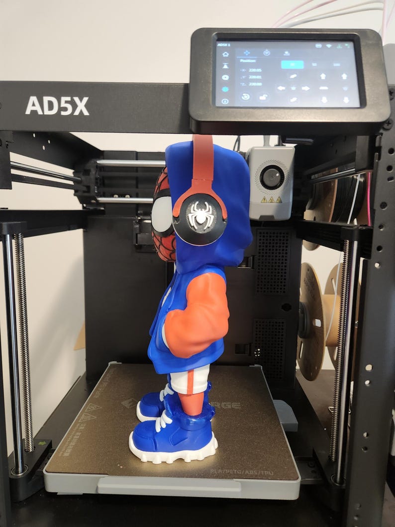 May include: A 3D-printed figurine of a superhero wearing a blue hoodie with orange sleeves, red headphones, and blue shoes. The figurine is on a 3D printer, with the model name "AD5X" visible.