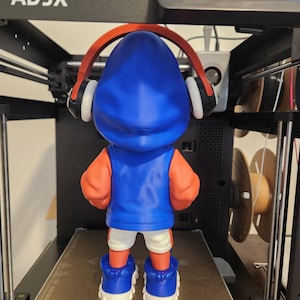May include: A 3D-printed figurine of a character wearing a blue hoodie, orange sleeves, white shorts, and blue sneakers. The figurine has red headphones and is standing on a 3D printer bed. The printer is black with the text "AD5X" on the top.