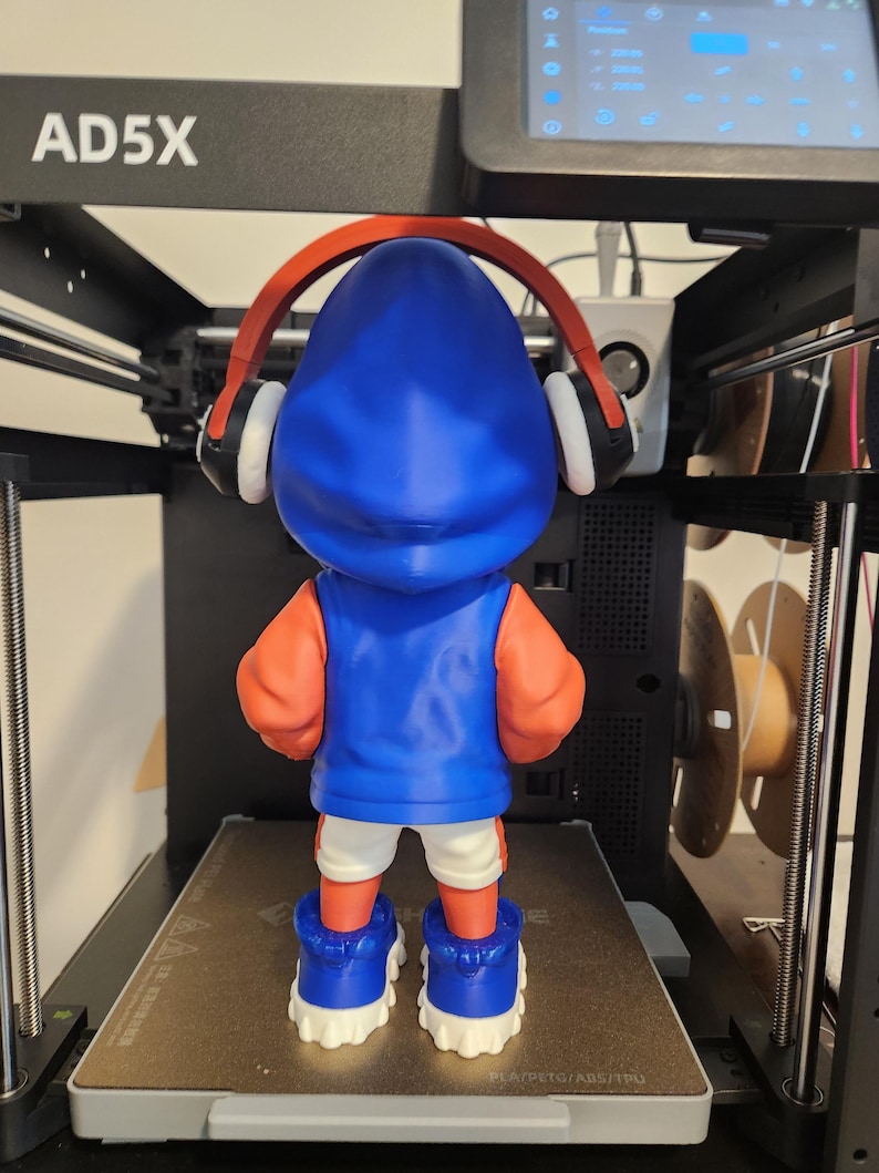 May include: A 3D-printed figurine of a character wearing a blue hoodie, orange sleeves, white shorts, and blue sneakers. The figurine has red headphones and is standing on a 3D printer bed. The printer is black with the text "AD5X" on the top.