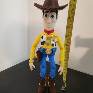 May include: A Sheriff Woody action figure wearing a brown cowboy hat, yellow long-sleeve shirt with black and white cow print vest, red neckerchief, blue trousers, and brown boots. A yellow measuring tape shows the figure is approximately 30 cm tall.