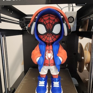 May include: A 3D-printed figurine of Spider-Man wearing a red and blue hoodie, white shorts, and blue sneakers. The figure has headphones and is standing on a 3D printer bed.