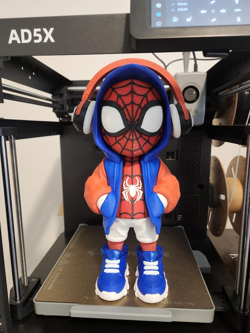 May include: A 3D-printed figurine of Spider-Man wearing a red and blue hoodie, white shorts, and blue sneakers. The figure has headphones and is standing on a 3D printer bed.