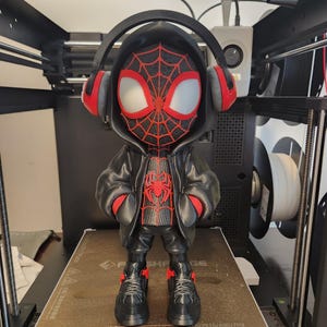 May include: A 3D-printed figurine of a superhero wearing a black hoodie and headphones. The figure has a red and black spiderweb design on the mask and chest. The figure is standing on a brown surface inside a 3D printer.