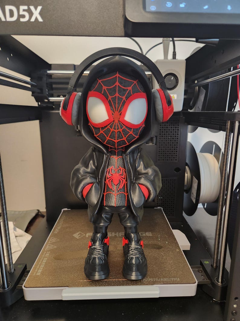 May include: A 3D-printed figurine of a superhero wearing a black hoodie and headphones. The figure has a red and black spiderweb design on the mask and chest. The figure is standing on a brown surface inside a 3D printer.