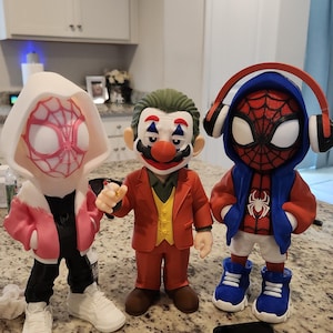 May include: Three figurines of comic book characters. One is a Spider-Woman in a white and pink hoodie, black pants, and white sneakers. The second is a Joker figure in a red suit and clown makeup. The third is a Spider-Man figure in a blue hoodie and red sneakers.