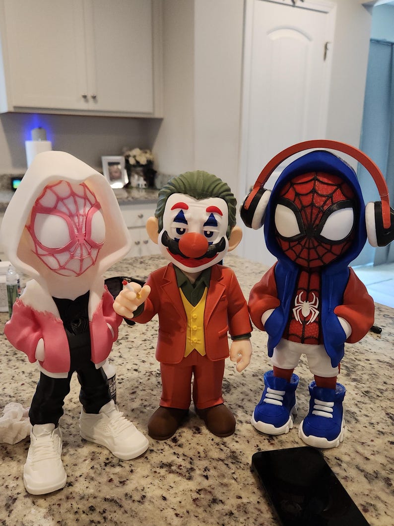 May include: Three figurines of comic book characters. One is a Spider-Woman in a white and pink hoodie, black pants, and white sneakers. The second is a Joker figure in a red suit and clown makeup. The third is a Spider-Man figure in a blue hoodie and red sneakers.