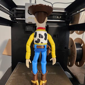 May include: A plastic action figure of a cowboy character standing on a 3D printer bed. The figure has a brown hat, a yellow long-sleeved shirt, a white vest with black cow-print spots, blue trousers, a brown belt with a holster, and brown boots.