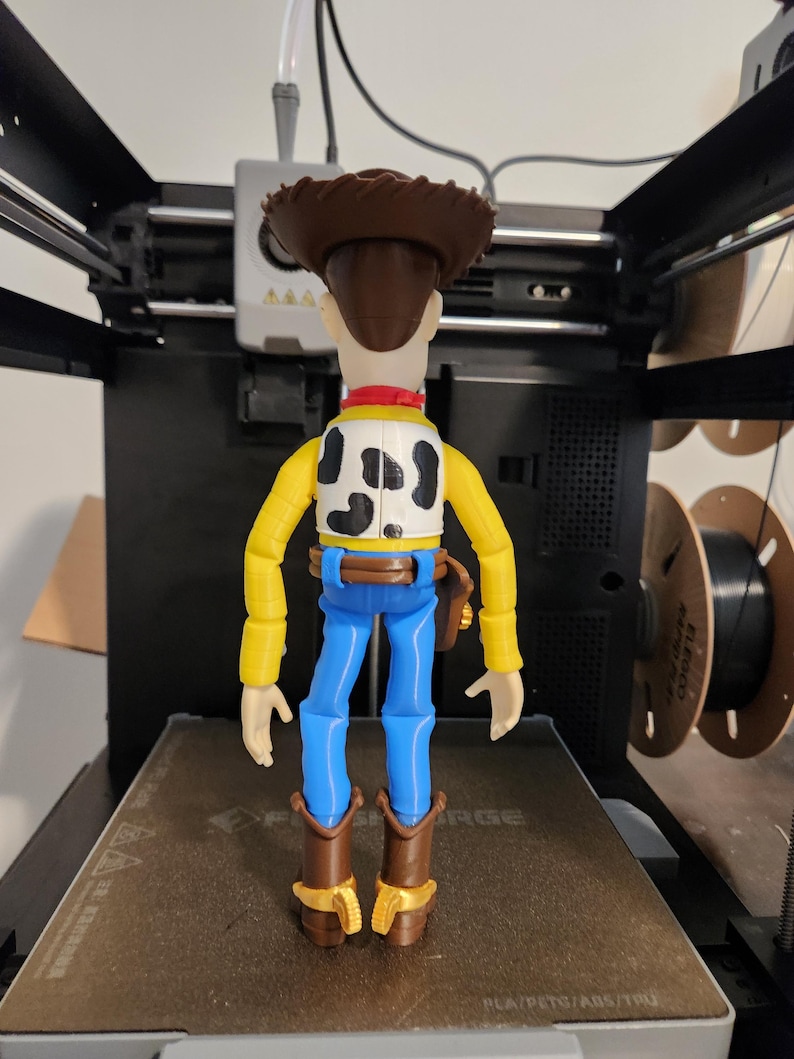 May include: A plastic action figure of a cowboy character standing on a 3D printer bed. The figure has a brown hat, a yellow long-sleeved shirt, a white vest with black cow-print spots, blue trousers, a brown belt with a holster, and brown boots.