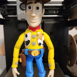 May include: A 3D printed action figure of a cowboy character wearing a brown hat, a yellow long-sleeved shirt with black and white cow-print vest panels, a gold sheriff badge, a blue belt with a gold buckle, blue trousers, and brown boots.