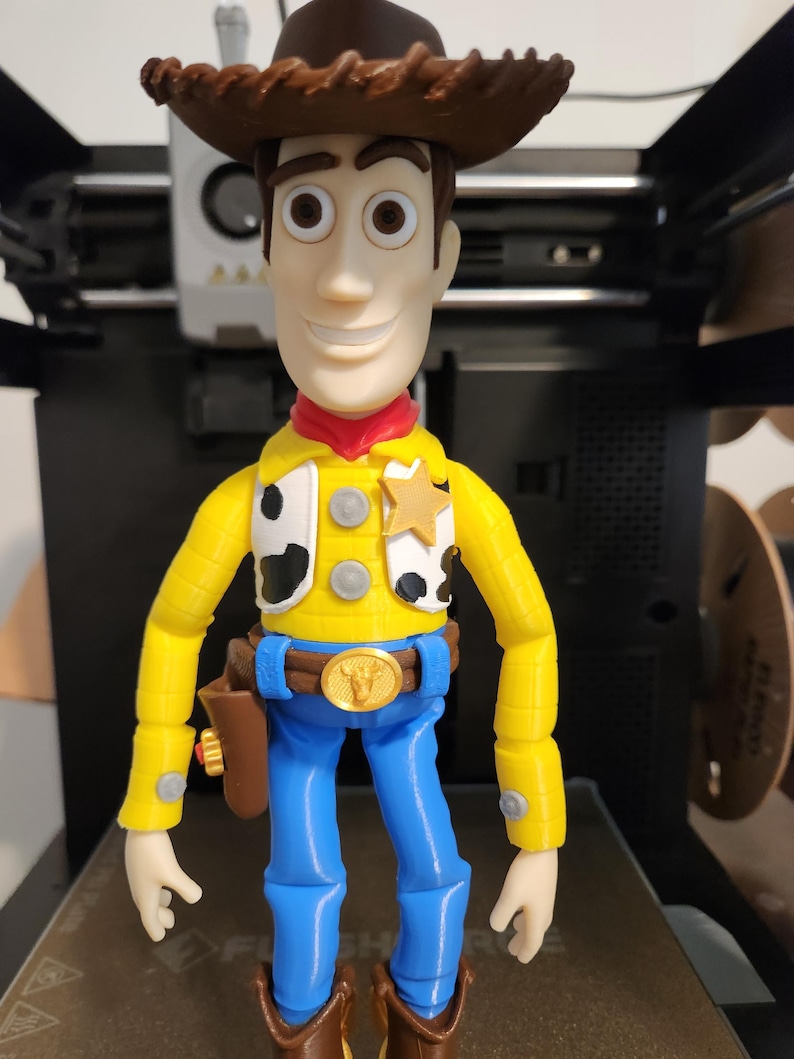 May include: A 3D printed action figure of a cowboy character wearing a brown hat, a yellow long-sleeved shirt with black and white cow-print vest panels, a gold sheriff badge, a blue belt with a gold buckle, blue trousers, and brown boots.