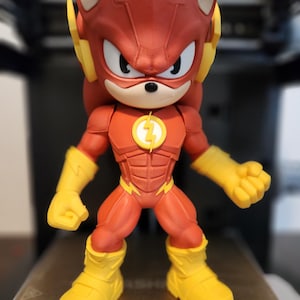 May include: A red and yellow action figure of a hedgehog-like character in a superhero costume. The figure has a red head with yellow ear pieces, a red and yellow suit with a lightning bolt emblem, and yellow gloves and boots. The figure is standing.