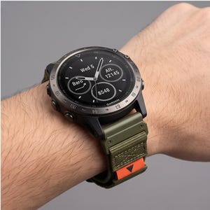 May include: A Garmin smartwatch with a round black digital display showing the date, altitude, barometric pressure, and step count. The watch features a dark metallic bezel and a textured olive green fabric strap with an orange accent tab.