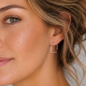 Op de afbeelding: Een kleine, dunne, zilverkleurige vierkante oorring gedragen in de oorlel. De oorbel heeft een minimalistische geometrische vorm met een eenvoudige draadconstructie, wat zorgt voor een strakke en ingetogen uitstraling bij het gezicht.