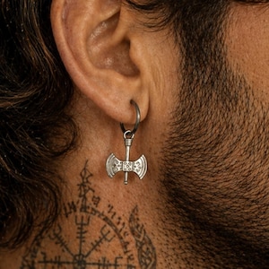 May include: A silver-colored earring in the shape of a double-bladed axe hangs from a hoop earring. The earring is worn on the ear. The person has a beard and a tattoo on their neck.