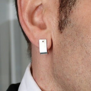 May include: A silver-toned hoop earring with a flat, rectangular front surface and a small, dark circular accent near the top. The earring is secured through the earlobe, featuring a smooth, metallic finish and a simple, clean-lined design.