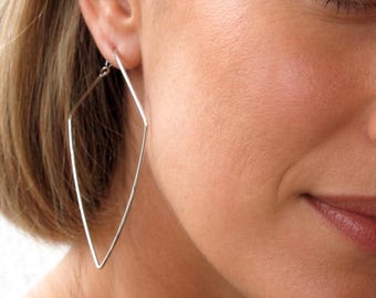 Puede incluir: Primer plano de una persona con pendientes geométricos plateados. Los pendientes tienen forma de diamante largo y delgado. La persona tiene el pelo rubio corto y lleva una camiseta negra.