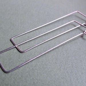 May include: A pair of silver-colored rectangular hoop earrings. The earrings are made of thin metal and have a simple, minimalist design. The hoops are long and slender, with a rectangular shape.