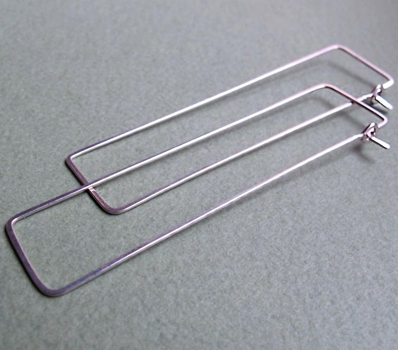 May include: A pair of silver-colored rectangular hoop earrings. The earrings are made of thin metal and have a simple, minimalist design. The hoops are long and slender, with a rectangular shape.