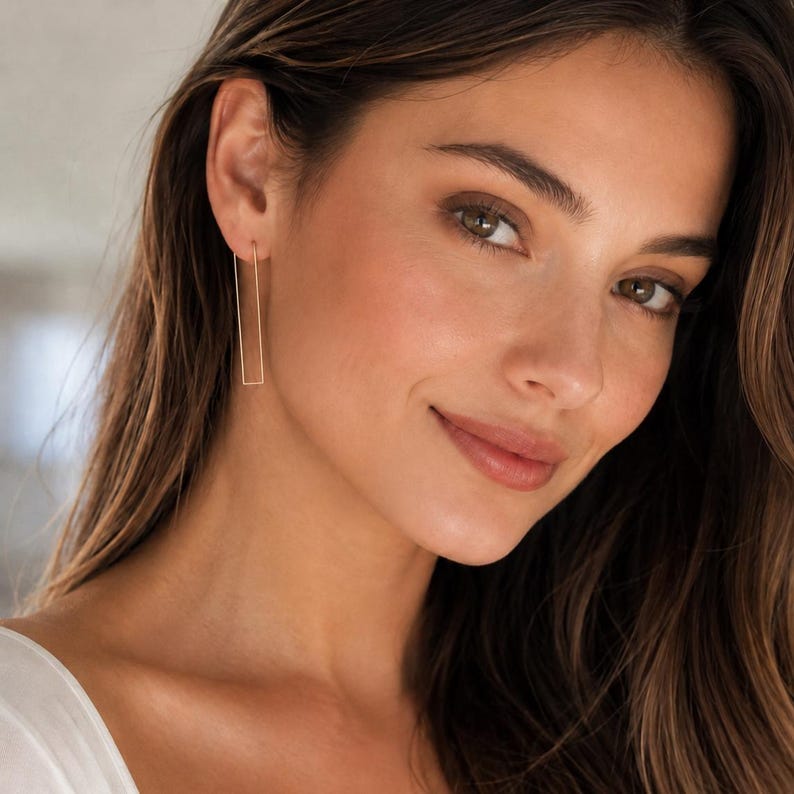 May include: Gold-colored rectangular drop earrings. The earrings are long and slender, with a simple, minimalist design. The earrings are shown on a person with brown hair and fair skin.